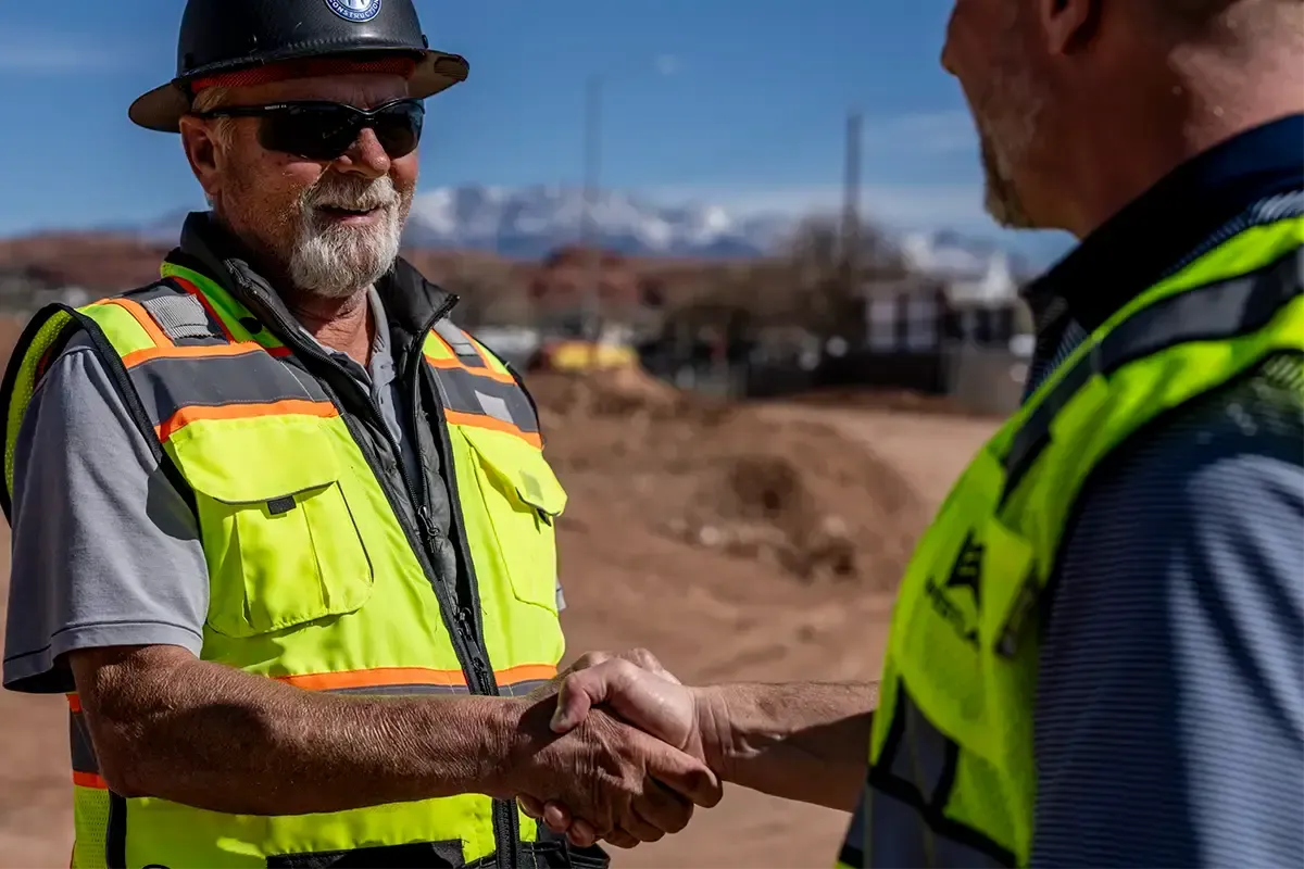 Two men in safety vests shaking hands on a job site with snow-capped mountains behind