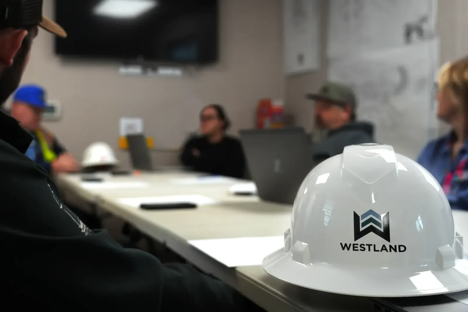 A white Westland hard hat on a conference table while a team meets around laptops
