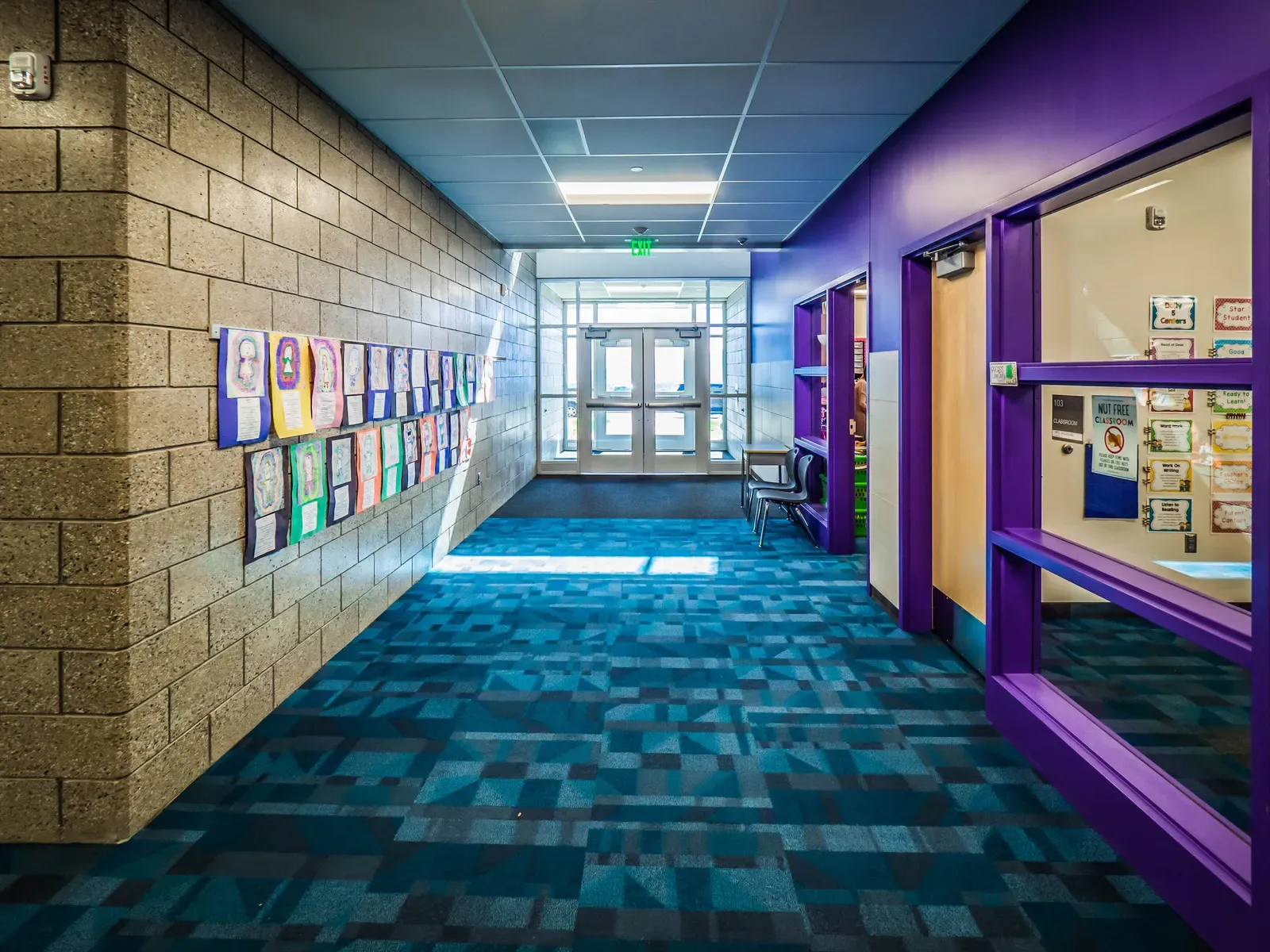 Corridor with a split-face block wall hung with student artwork, a purple-framed classroom window and blue patterned carpet