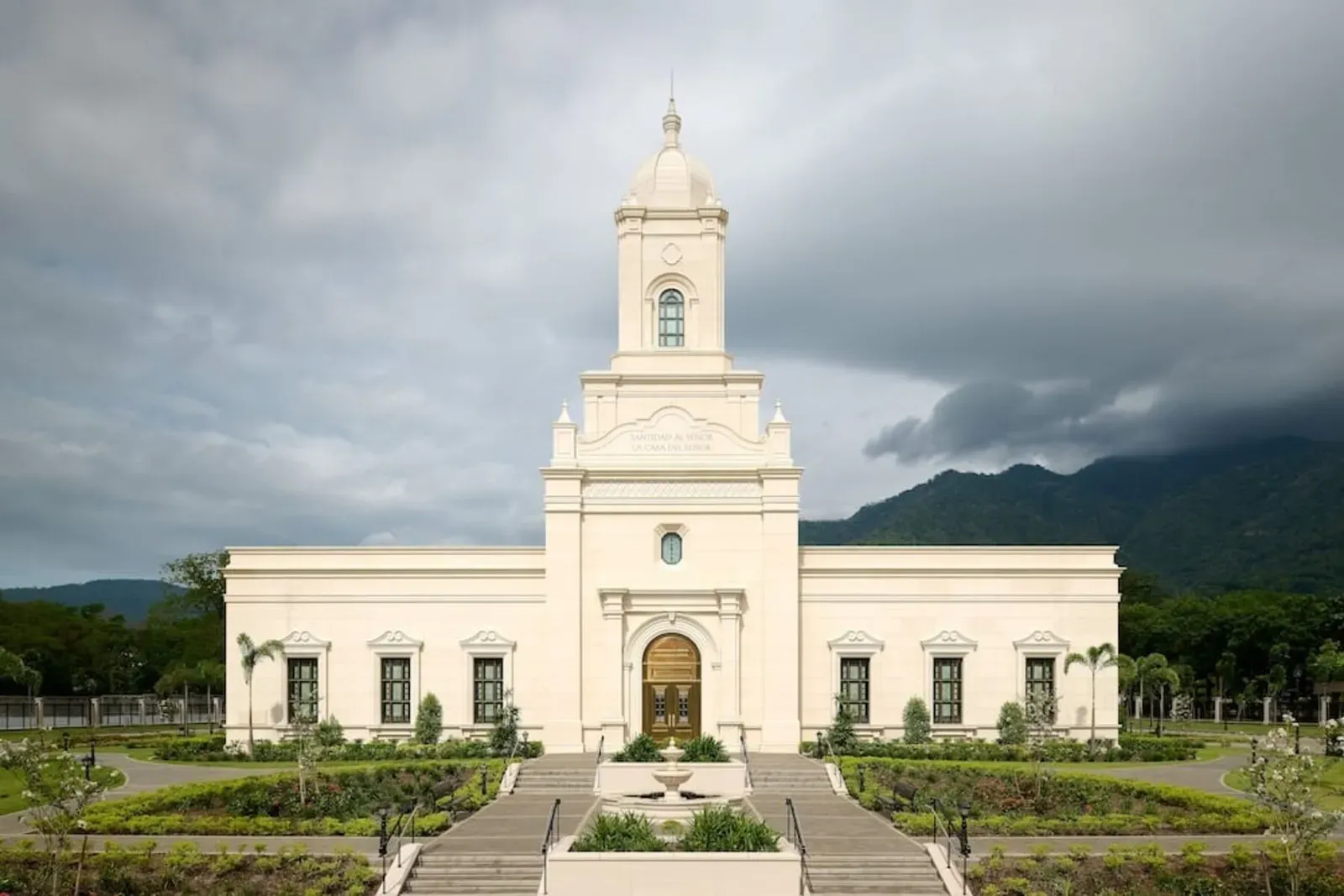 Front of the cream-colored temple with its domed central tower, a fountain and stairs, under a stormy sky with mountains behind