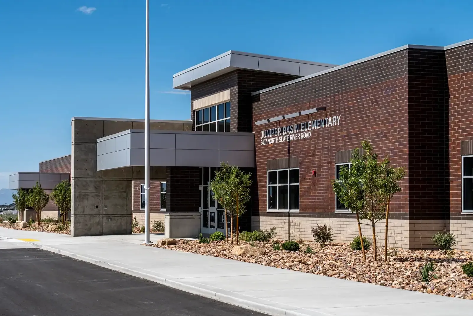 Angled view of the school entrance with a silver canopy, a concrete wall, brick walls and young trees in a rock landscaped bed.
