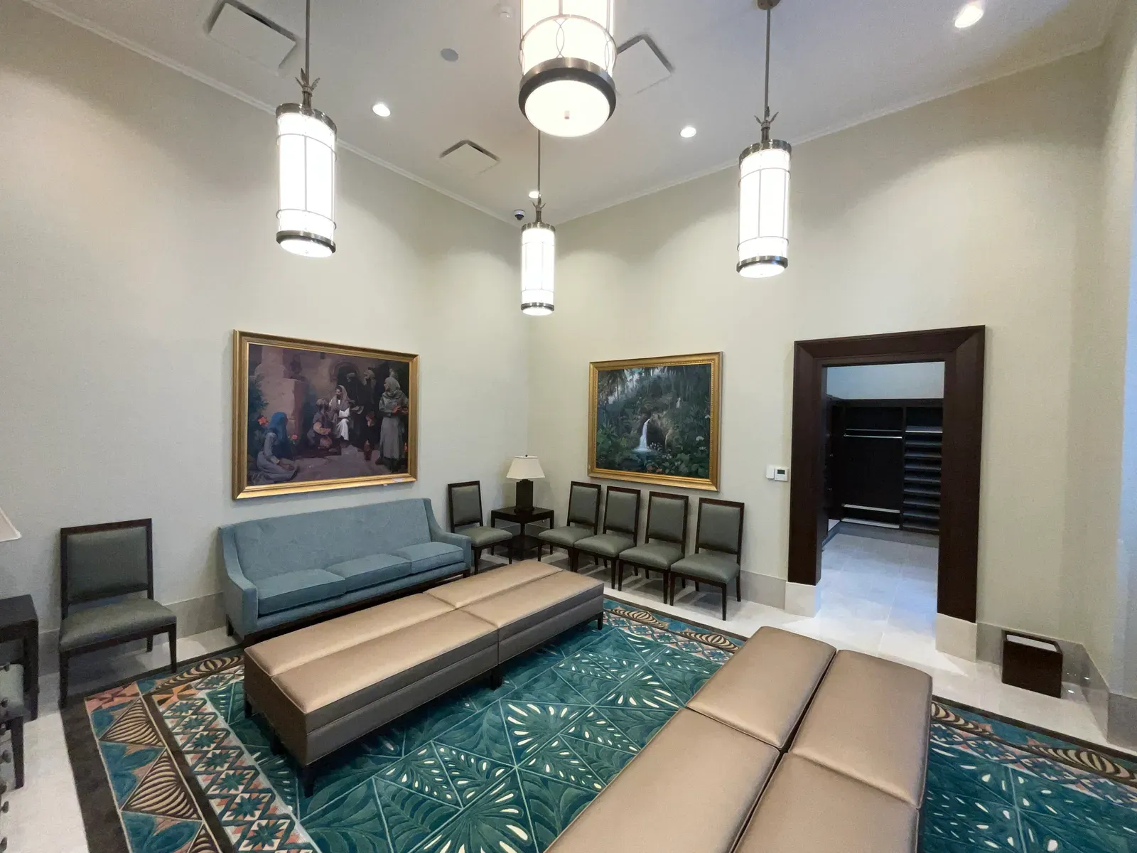 Sitting room with a blue sofa, tan ottomans, framed paintings and a teal rug with leaf and geometric patterns.