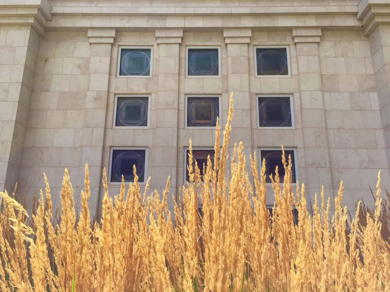 Stone exterior wall with rows of small art-glass windows, partly hidden behind tall golden grass.
