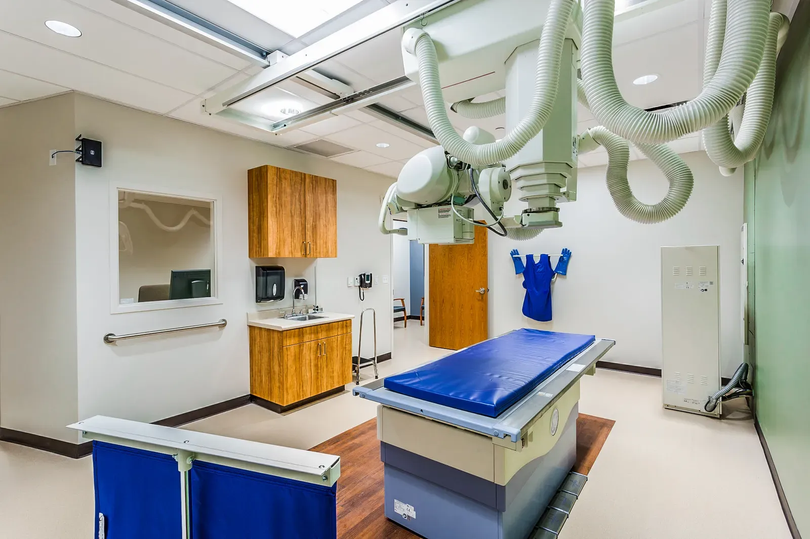 X-ray room with a ceiling-mounted x-ray unit, a padded table, a lead-glass viewing window and wood cabinetry