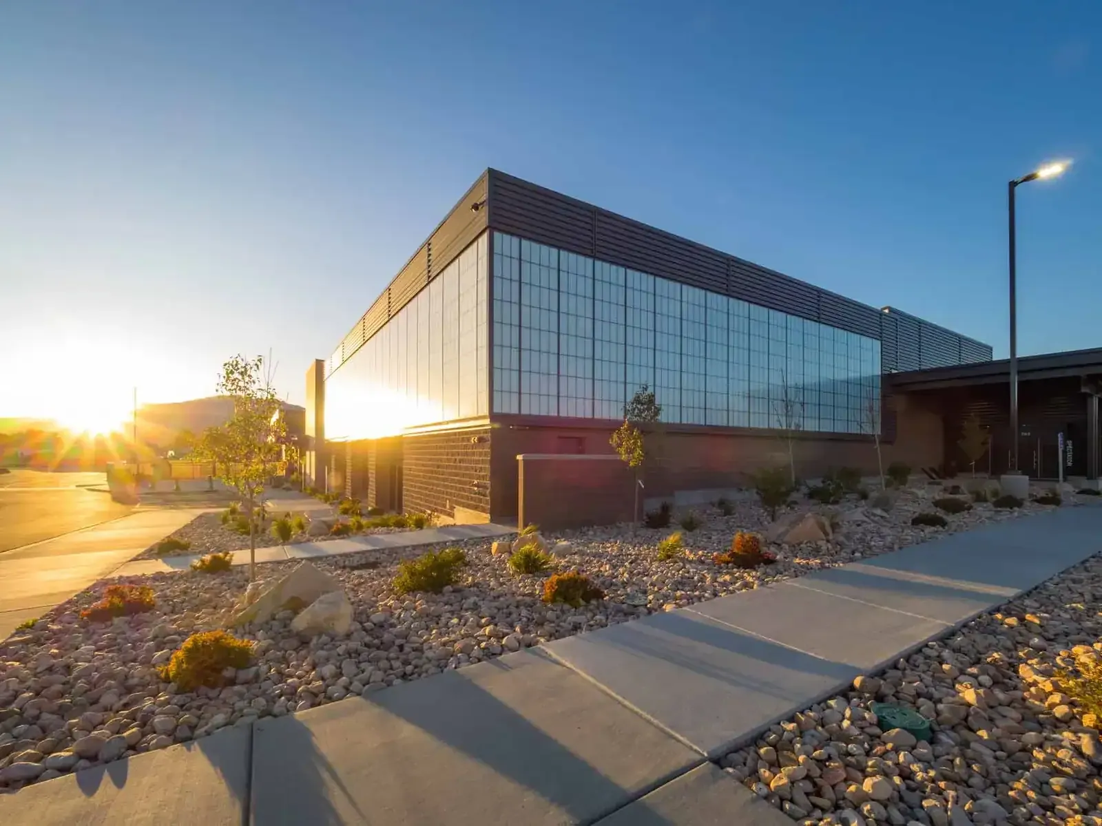 Corner of the aquatic center at sunset with a tall translucent glass-panel wall above a brick base and rock landscaping.