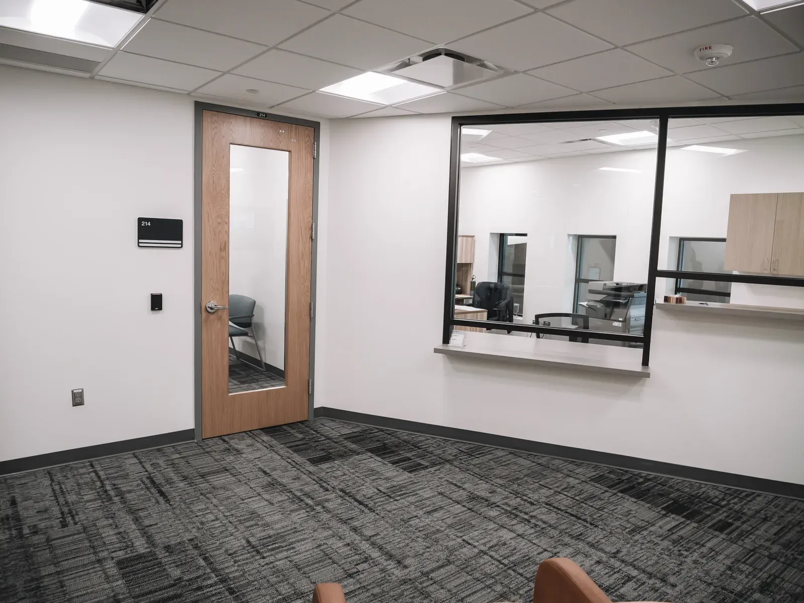 Waiting area with a wood-framed glass door beside a service window with a counter, on gray patterned carpet.
