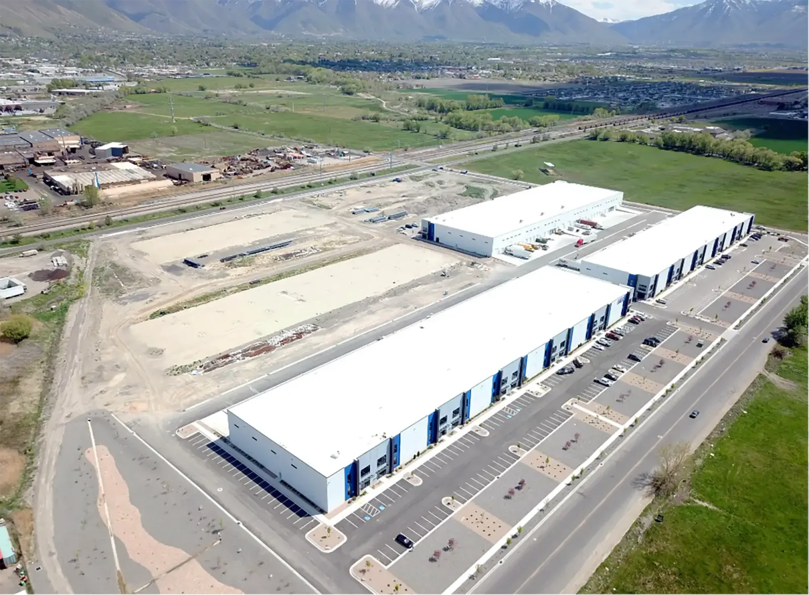 Aerial view of several white-roofed industrial buildings with parking lots and open ground beside them.