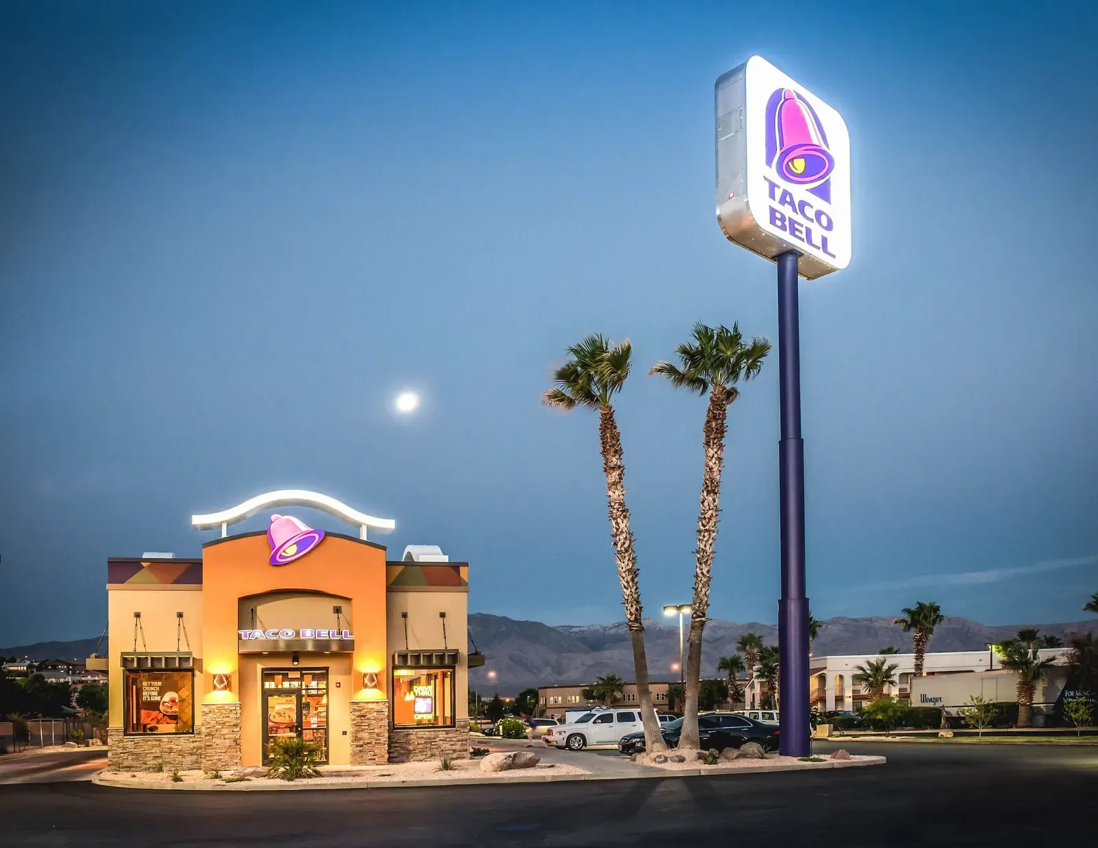 Taco Bell at dusk with palm trees, a tall pylon sign and mountains in the background.