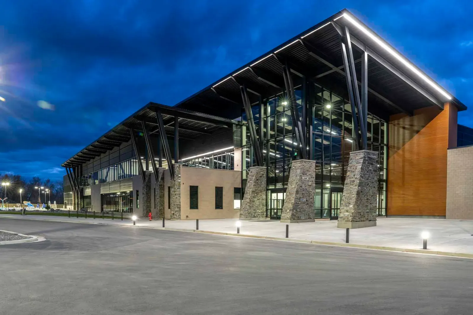 Entrance side of the recreation center at dusk, with angled steel columns, stone piers, a glass curtain wall and a wood-clad corner.