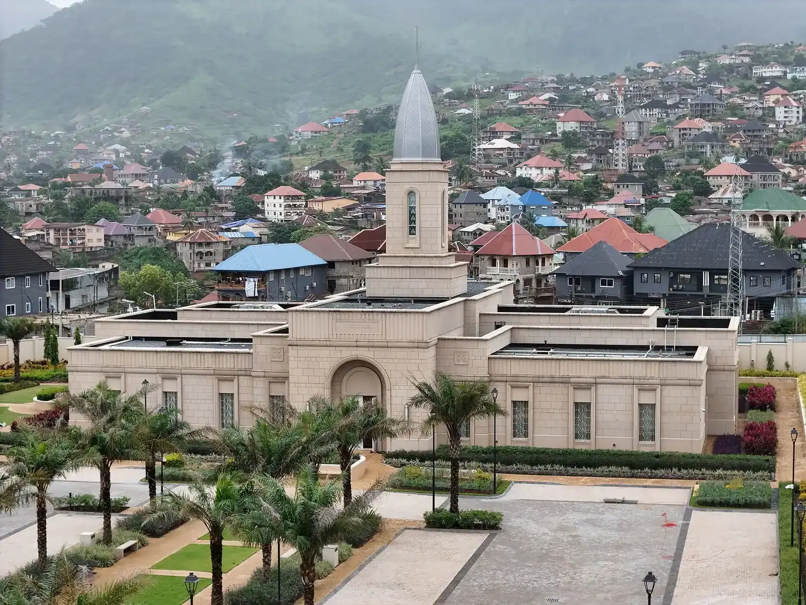 Aerial view of the temple's stone walls and gray spire above a courtyard planted with palms, with hillside houses behind.