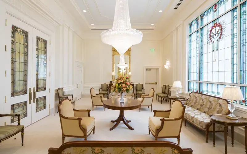 Room with a tall crystal chandelier, a round table with flowers, armchairs, a settee and a large stained-glass window with a wreath design