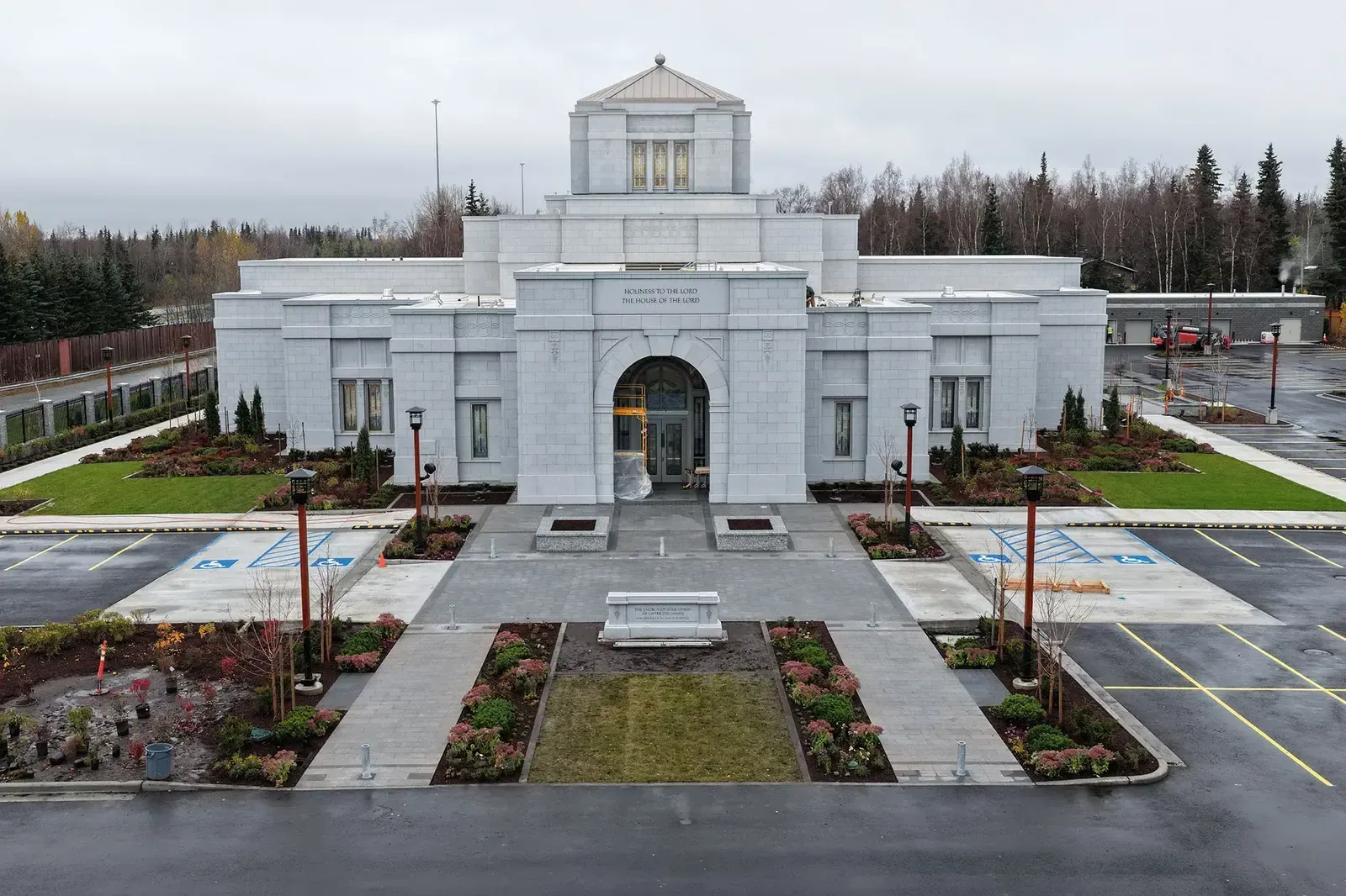 Front of the temple with a central arched entrance, an inscription over the door, a stone walkway with new landscaping and spruce trees behind.