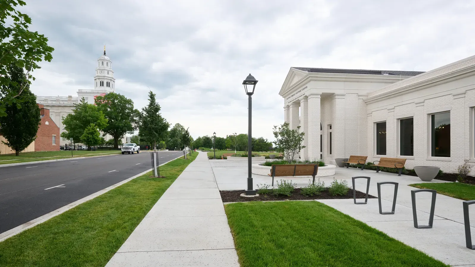 White brick visitors’ center with columns and wood benches beside a sidewalk, with the temple’s white tower in the distance down the street.