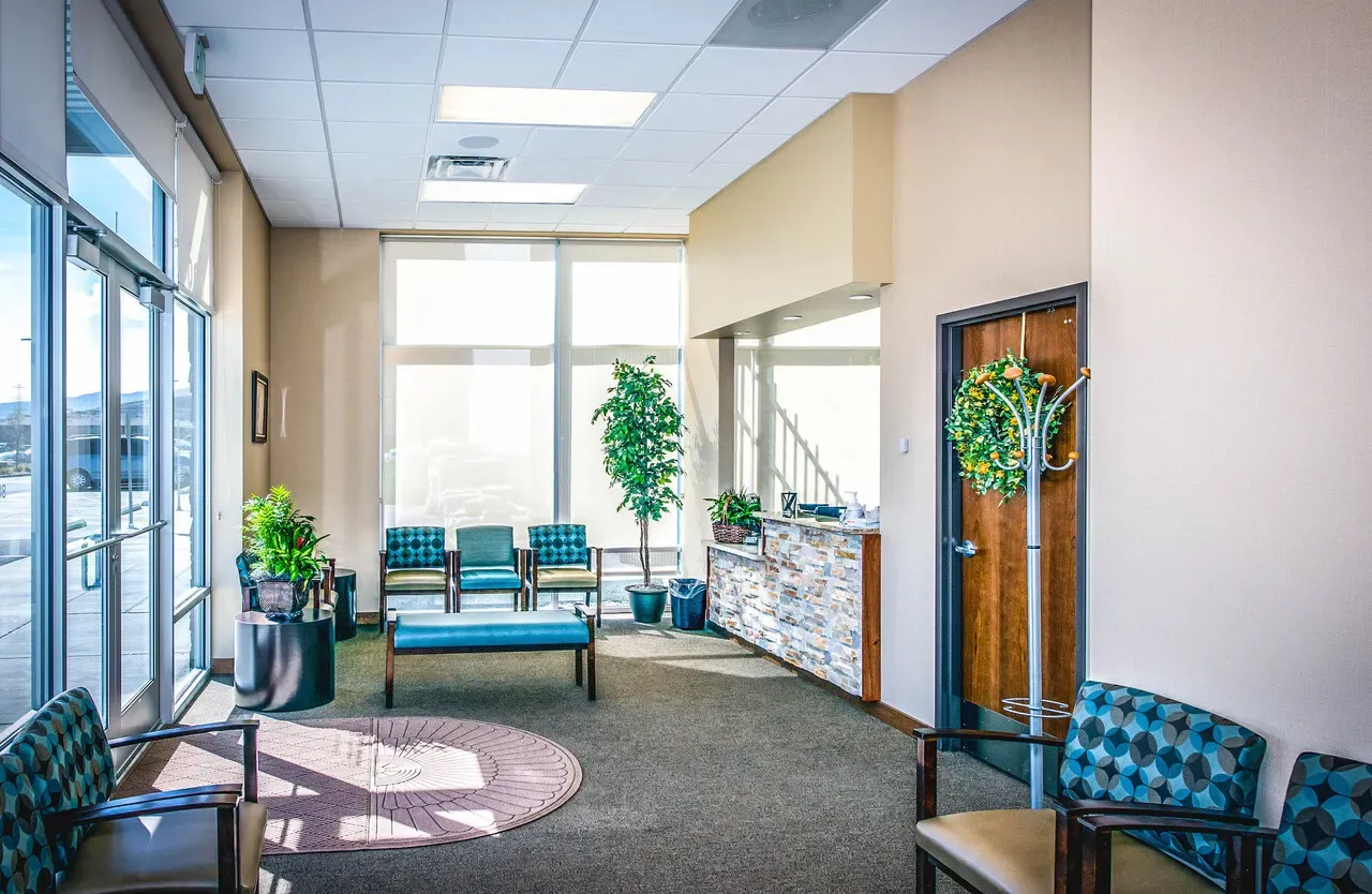 Waiting room with teal chairs, a bench and plants beside a large window wall.