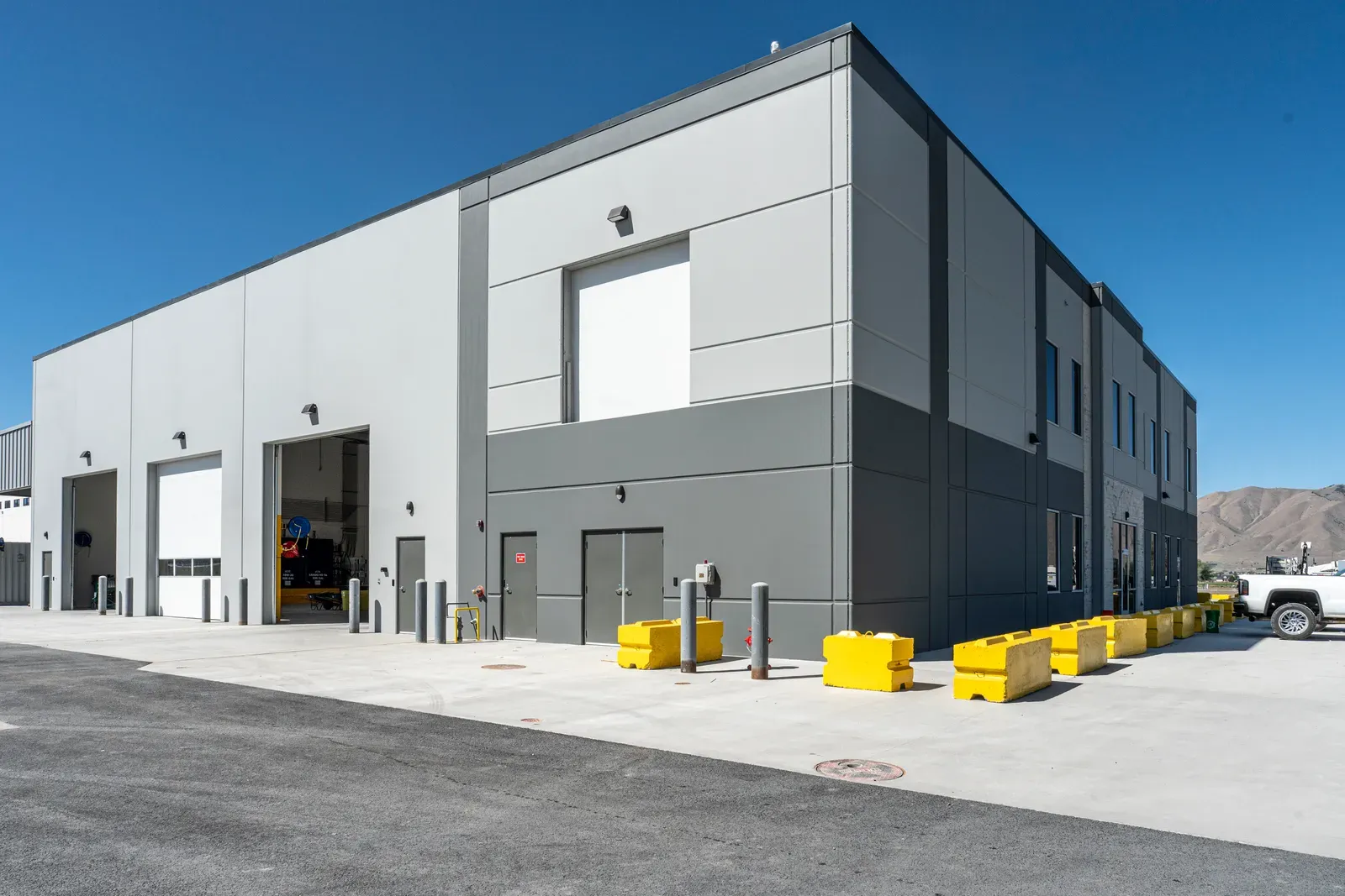 Exterior of the gray and white tilt-up building with overhead doors, a two-story section and yellow barriers.