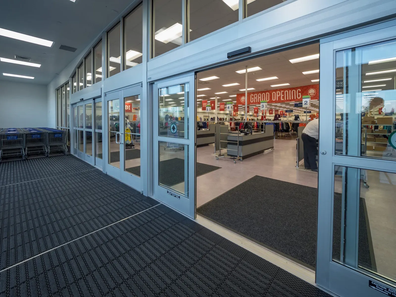 Glass entry doors and floor mats looking into the store, with a grand opening banner and checkout counters inside.