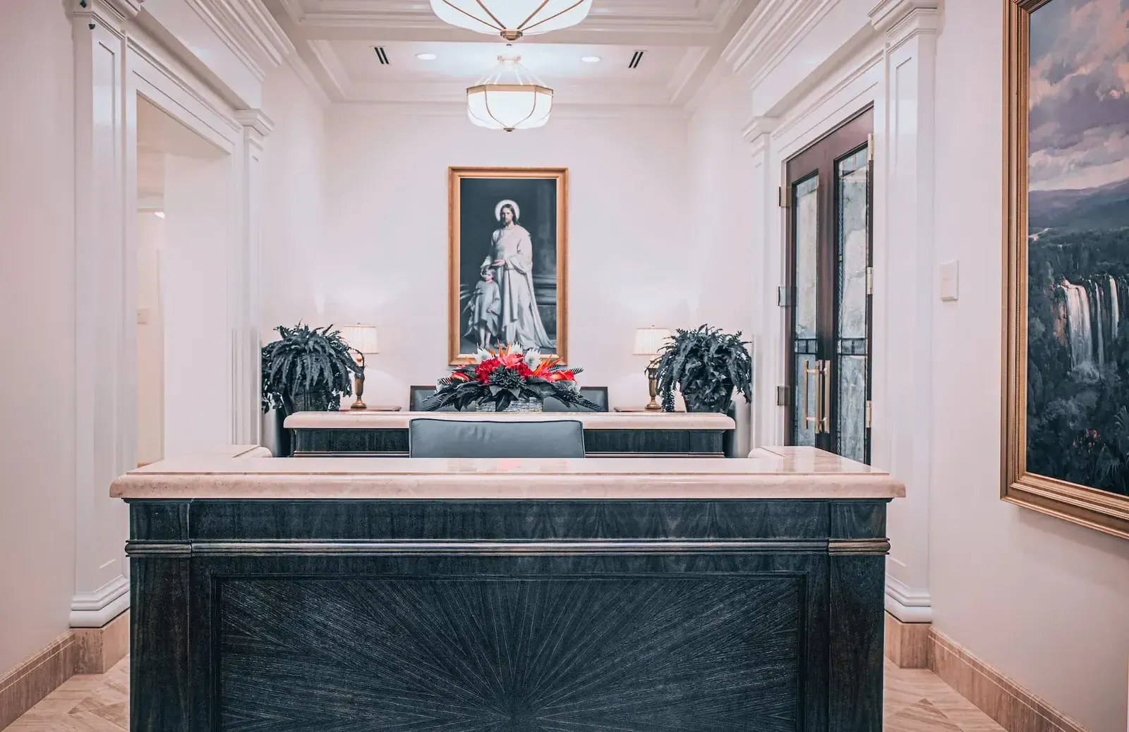 Reception desk in a white paneled lobby, with a framed religious painting behind it and a waterfall landscape on the right wall.