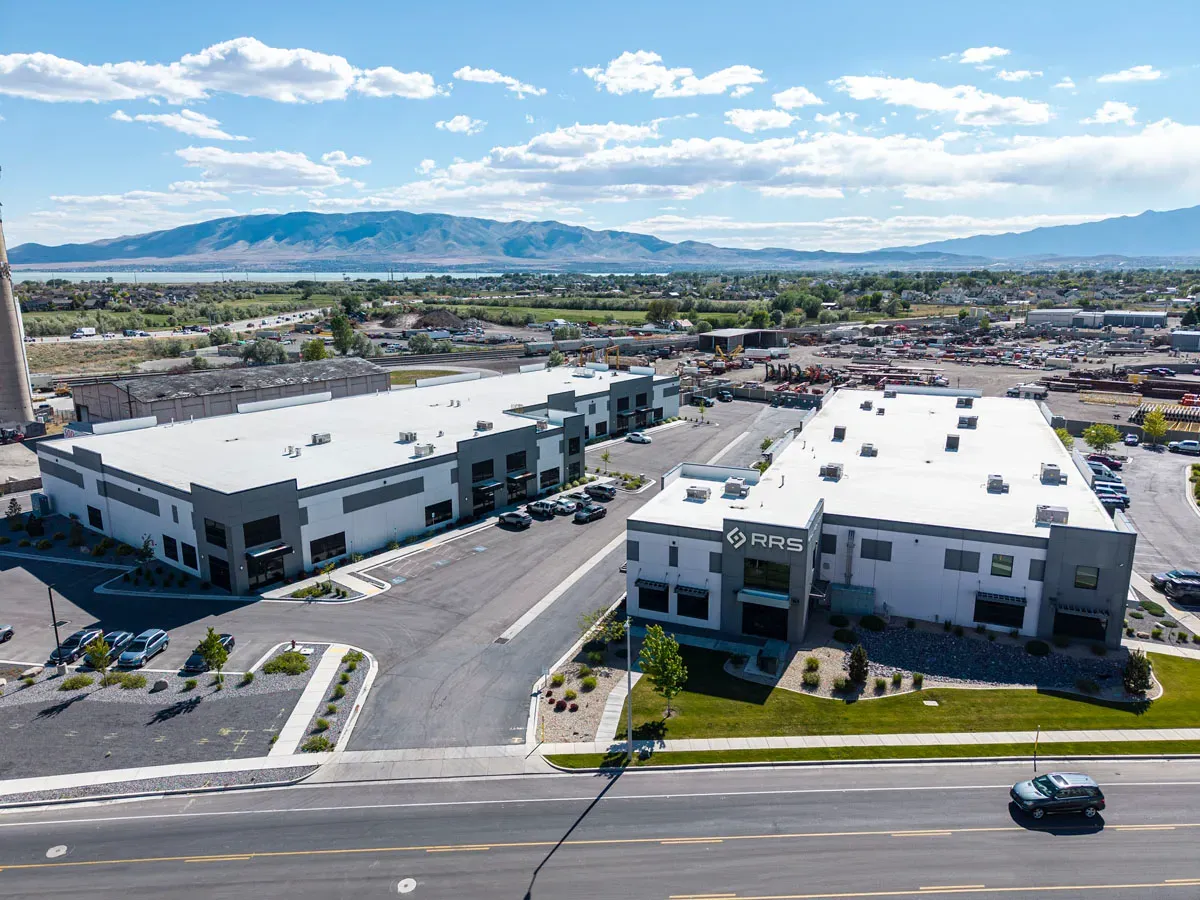 Aerial view of two industrial buildings with flat white roofs, a parking lot between them and mountains in the distance.