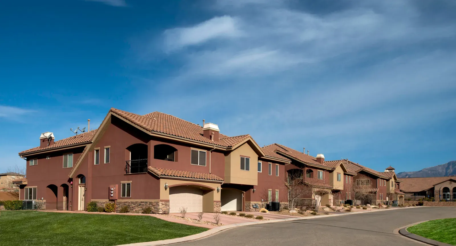 Row of two-story condominiums in red and tan stucco with tile roofs and garages along a curving street, with mountains in the distance.