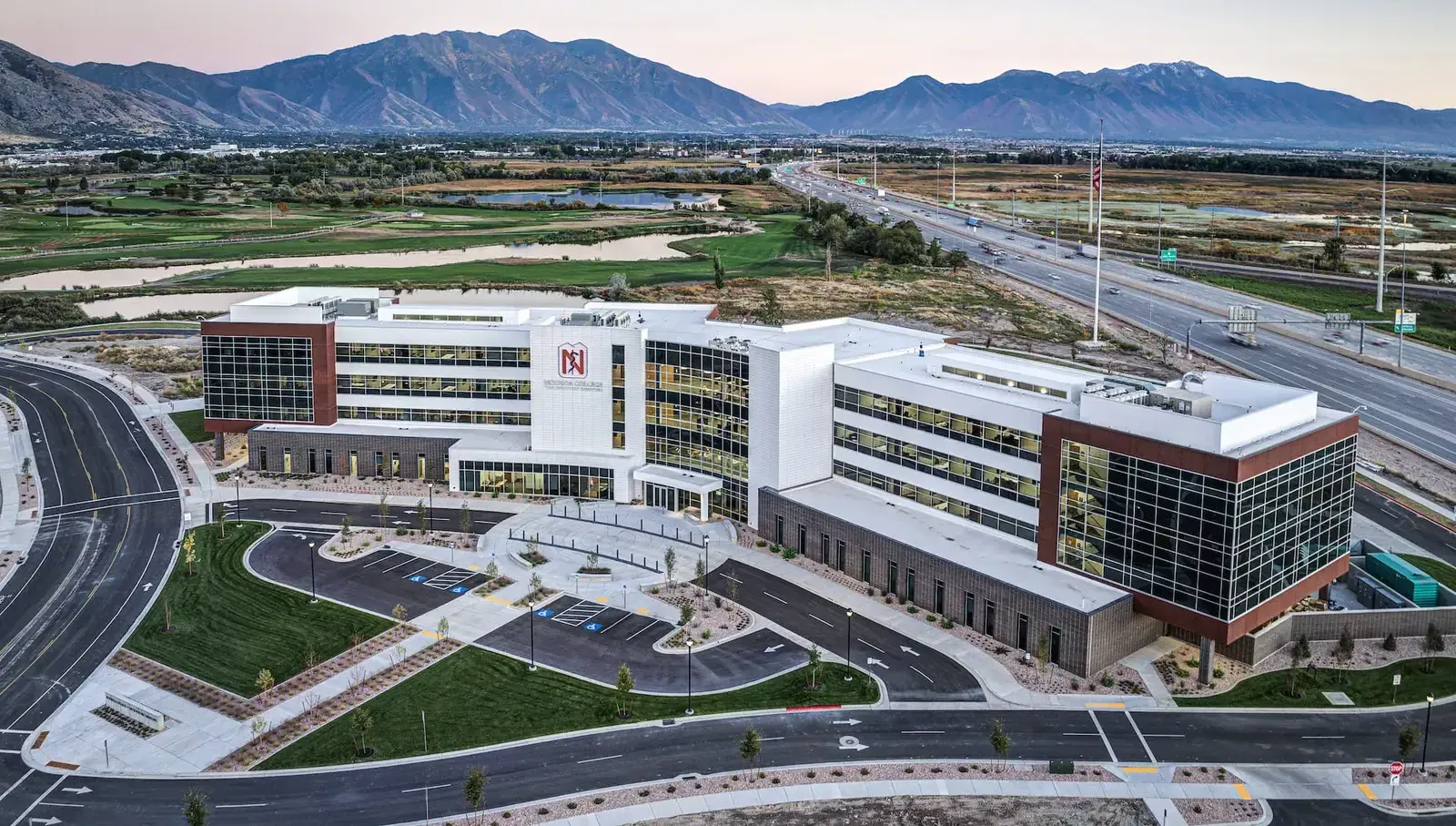 Aerial view of the multi-story college building and its parking lots, with mountains in the distance.