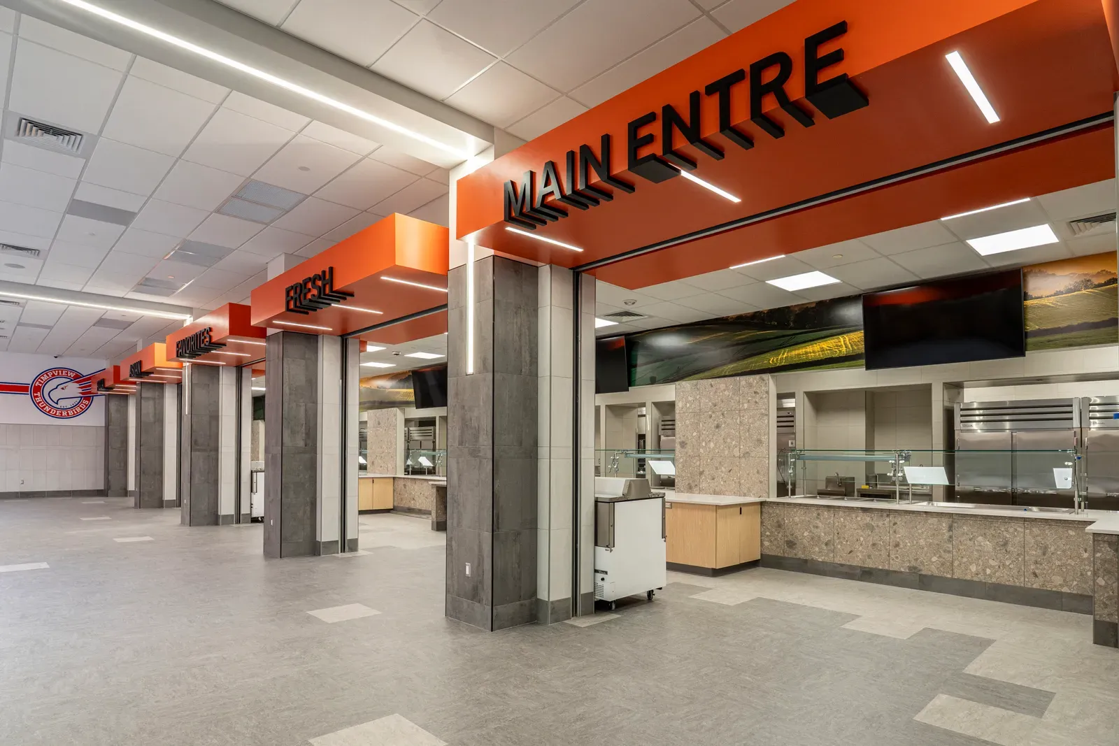 Cafeteria serving counters with orange signs reading Main Entre and Fresh above gray stone-clad columns.