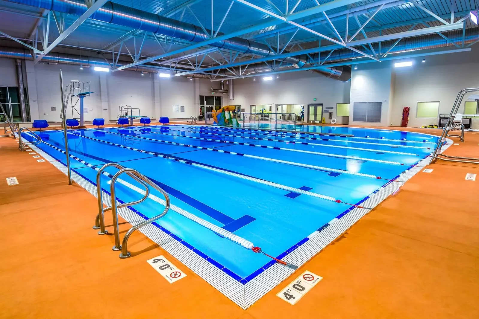 Indoor lap pool with marked lanes, a diving board, and an orange deck under a steel-truss roof