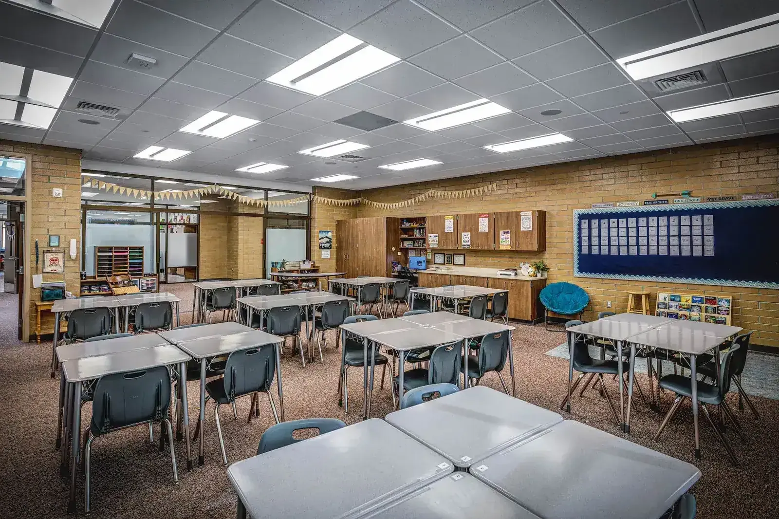 Classroom with groups of gray desks and chairs, a brick wall with a bulletin board, and a glass wall opening to the room beyond.