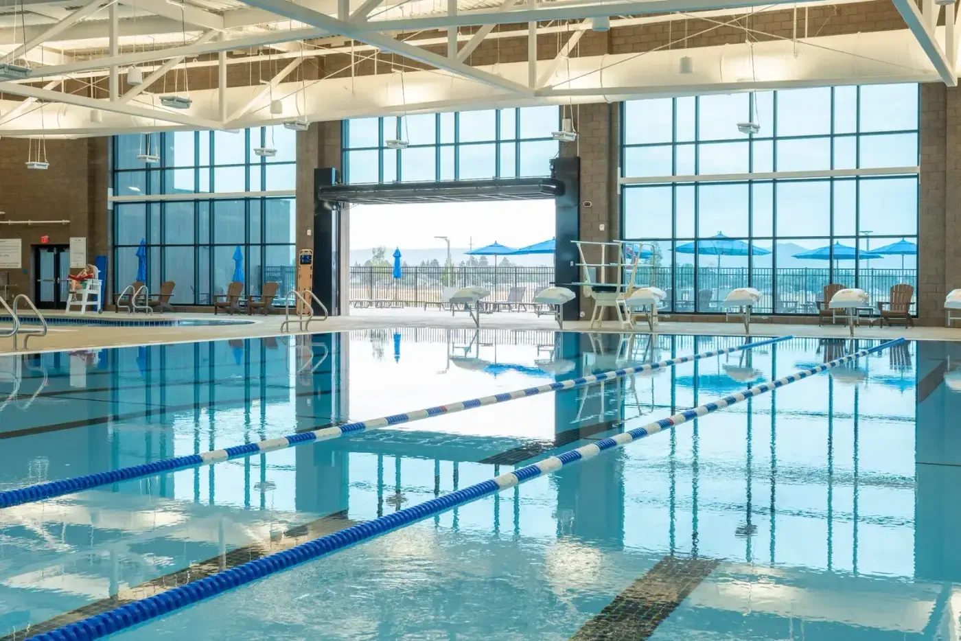 Pool with blue and white lane lines in the foreground, a lifeguard stand and tall windows, with a patio visible through an open overhead door.