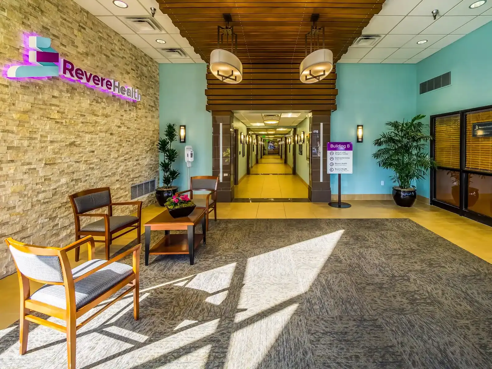 Lobby with a stone feature wall and illuminated Revere Health sign, wood-framed chairs and a long corridor leading away.