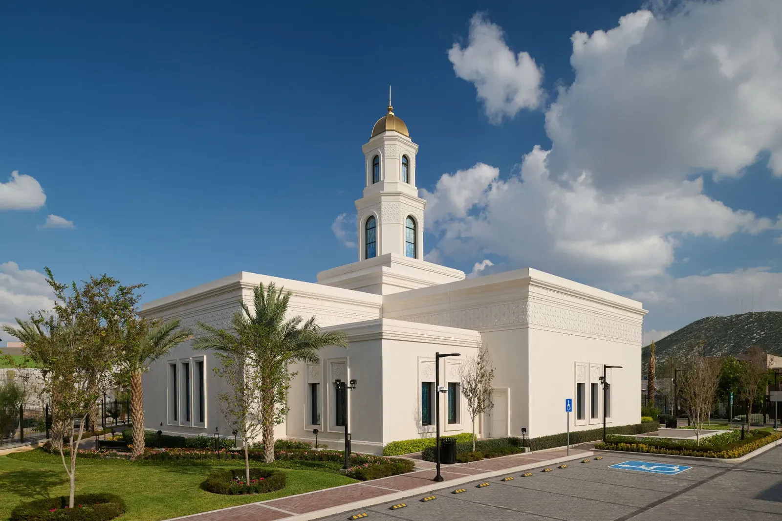 Side of the white temple with its tower and gold dome, palm trees and a landscaped lawn beneath a cloudy blue sky.