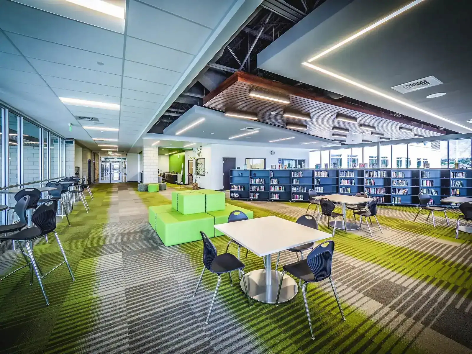 Library and commons with green seating blocks, blue bookshelves and striped green carpet beneath a wood-slat ceiling