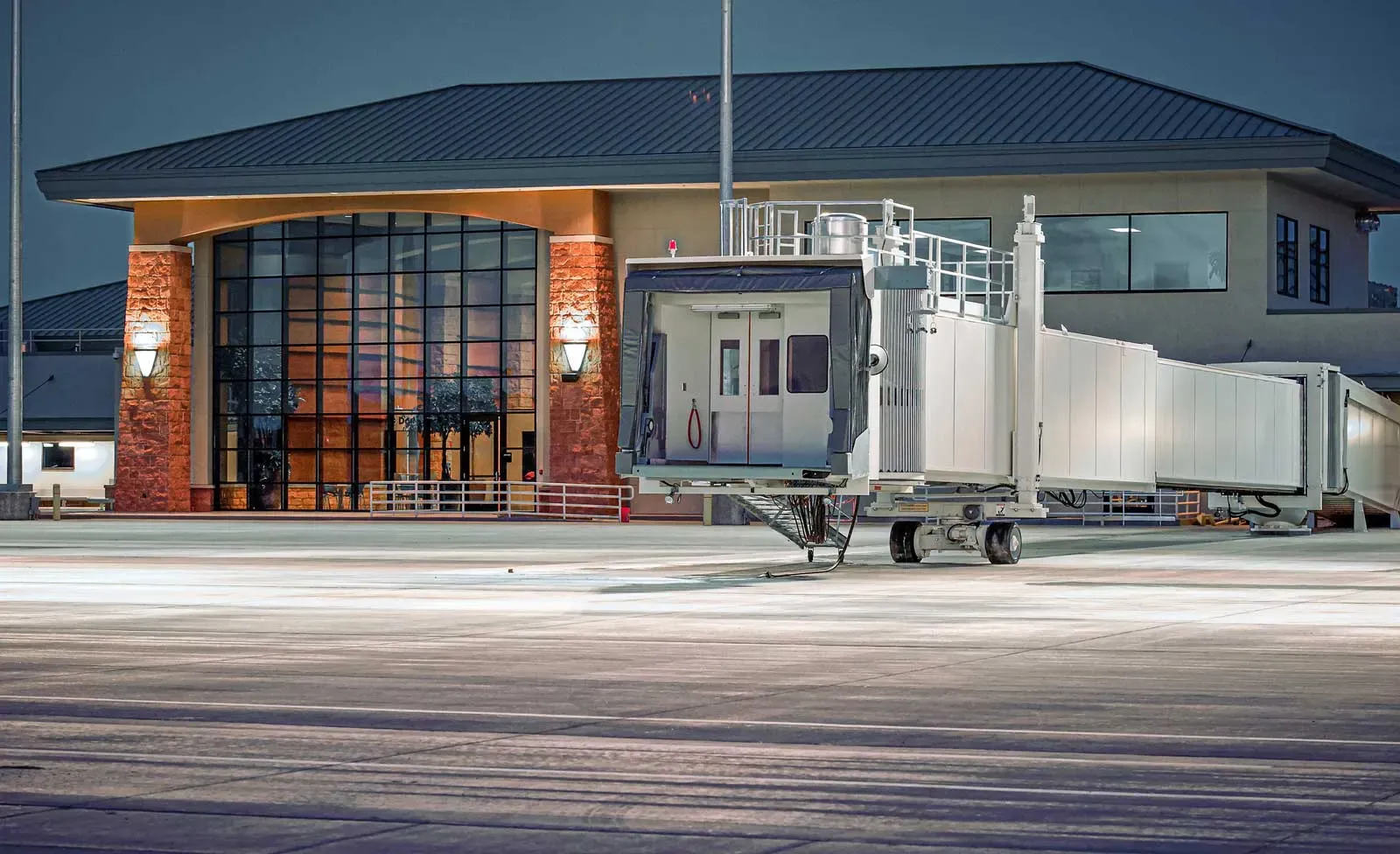 Terminal exterior at dusk with a glass-fronted entry framed by red stone piers, and a passenger boarding bridge extended at right.