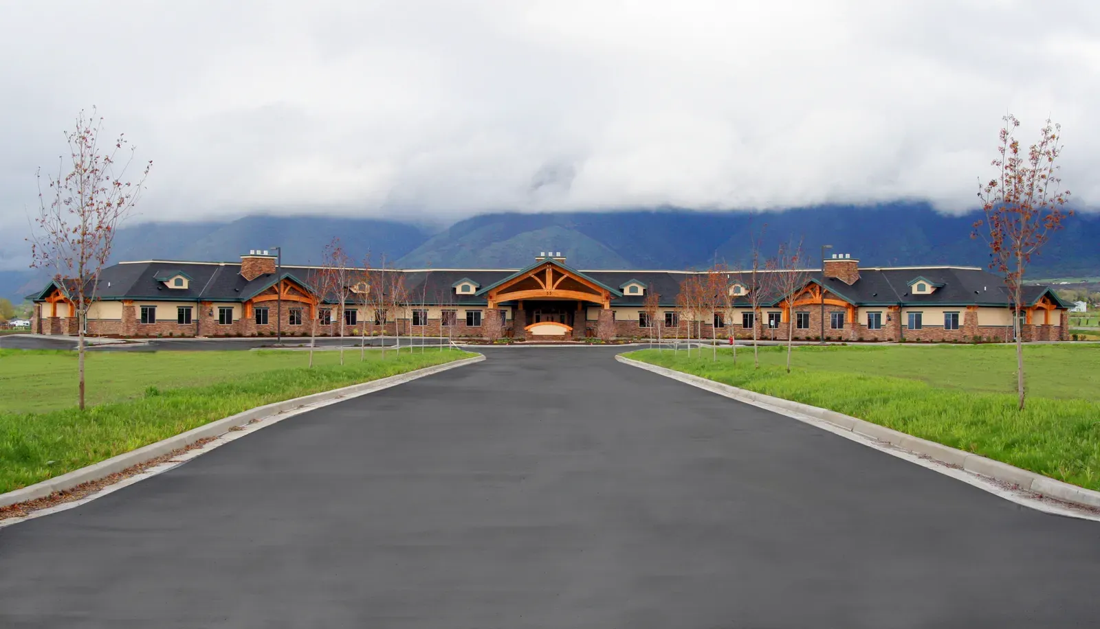 Front view of the long, low building with a central timber entry and mountains behind a cloudy sky.