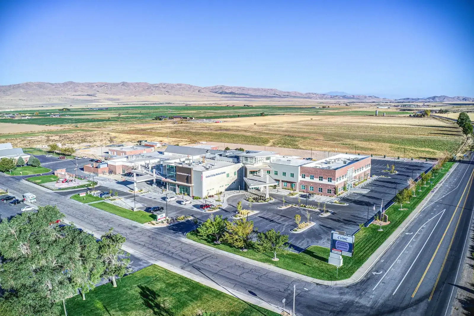 Aerial view of the hospital campus and parking lots beside a road, with farmland and hills beyond