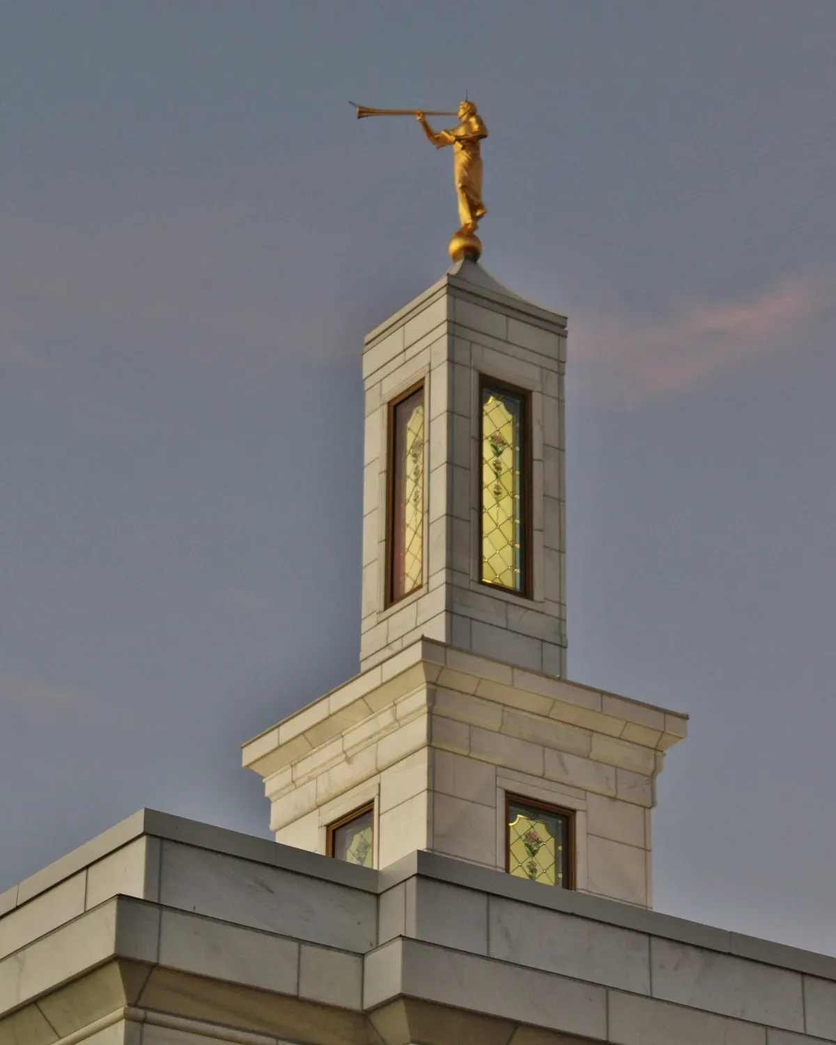 Front of the temple in evening light with an illuminated entrance sign, a black iron fence, autumn flower beds and the spire against a pink sky.