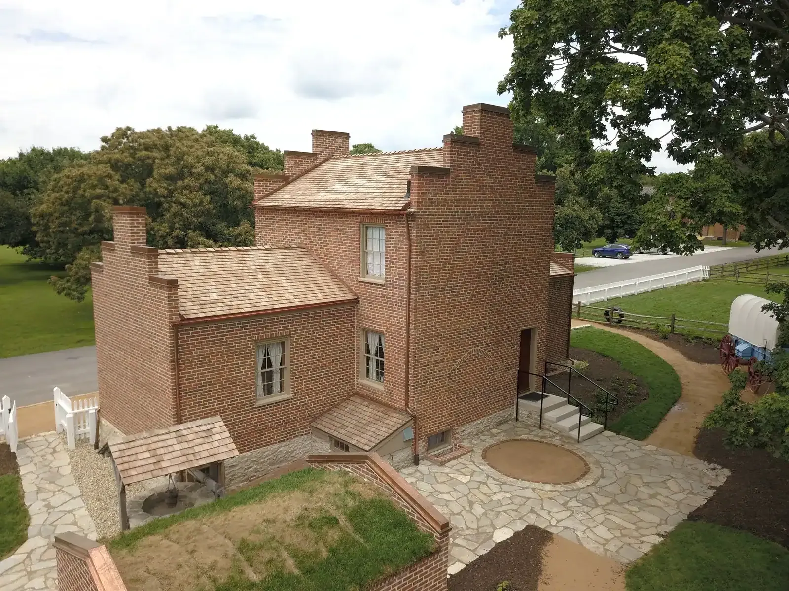 Aerial view of the back of the brick home with its stepped gables, a stone patio, a small shingle-roofed structure and a covered wagon at the edge of the lawn.