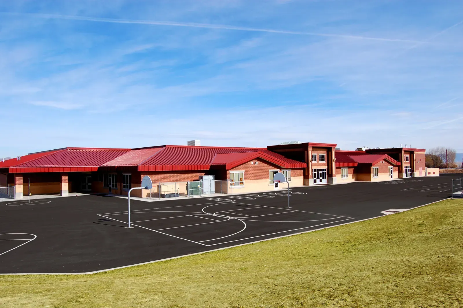 Long, low brick school with red metal roofs beside an asphalt playground with basketball court lines and a grass field.