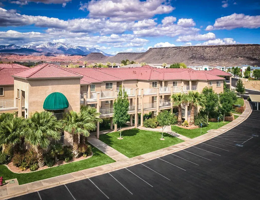 Aerial view of a three-story stucco resort building with red tile roofs, a green awning and lawns, with mountains in the distance