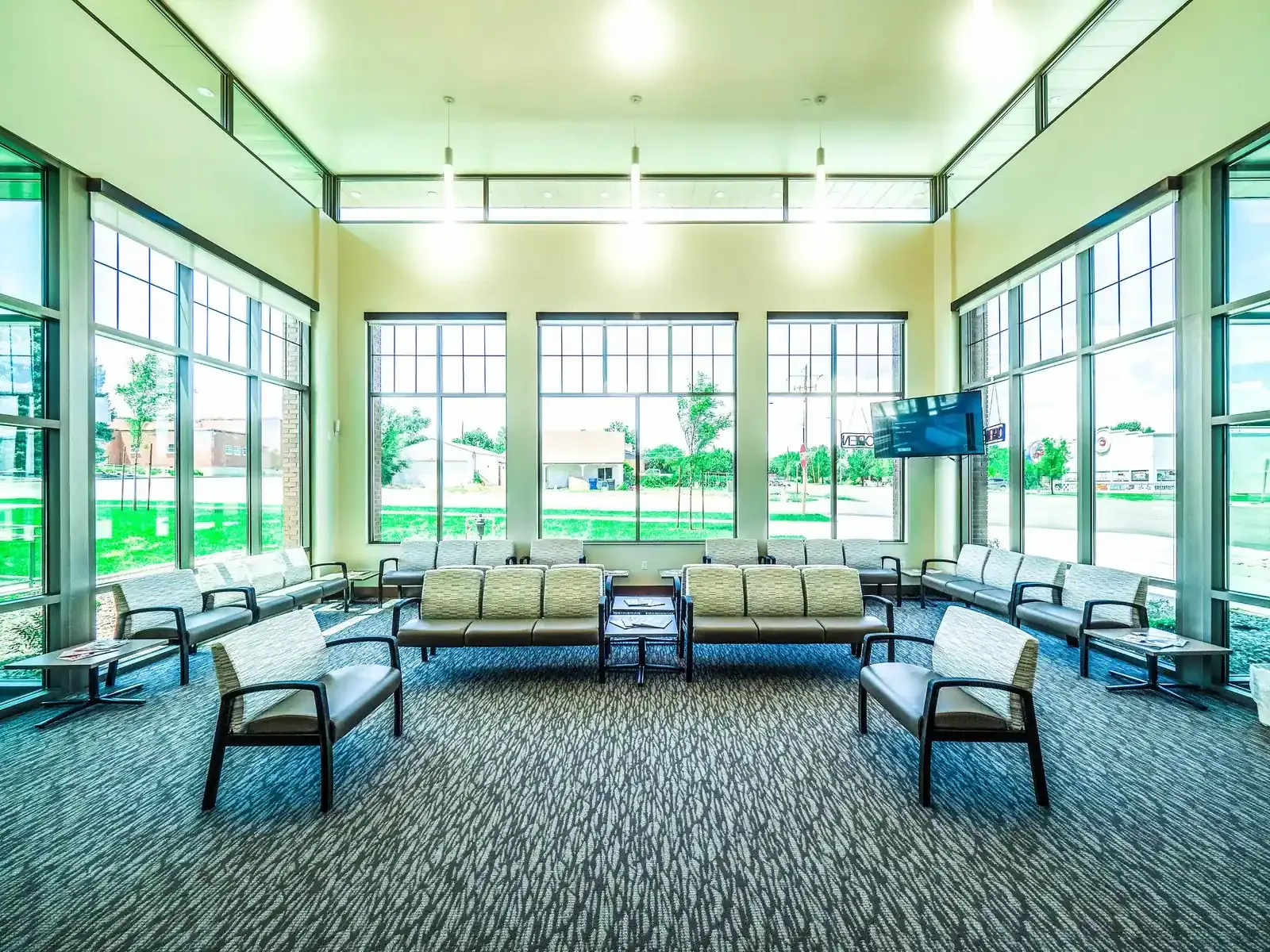 Waiting area with rows of seating facing tall windows and a wall-mounted TV.
