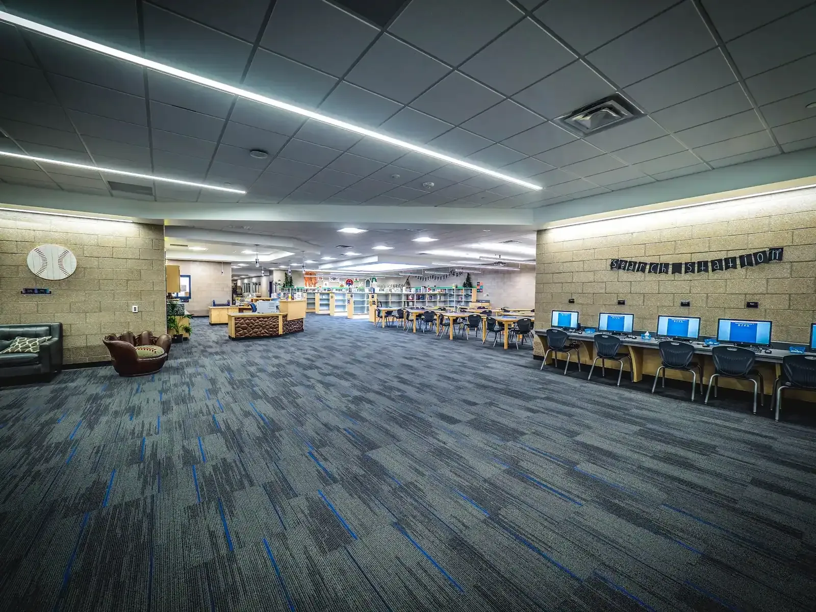 Library with bookshelves, a row of computer stations, and lounge seating under long light fixtures