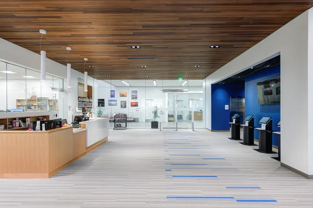 Library lobby with a wood ceiling, a curved service desk and self-checkout kiosks against a blue wall