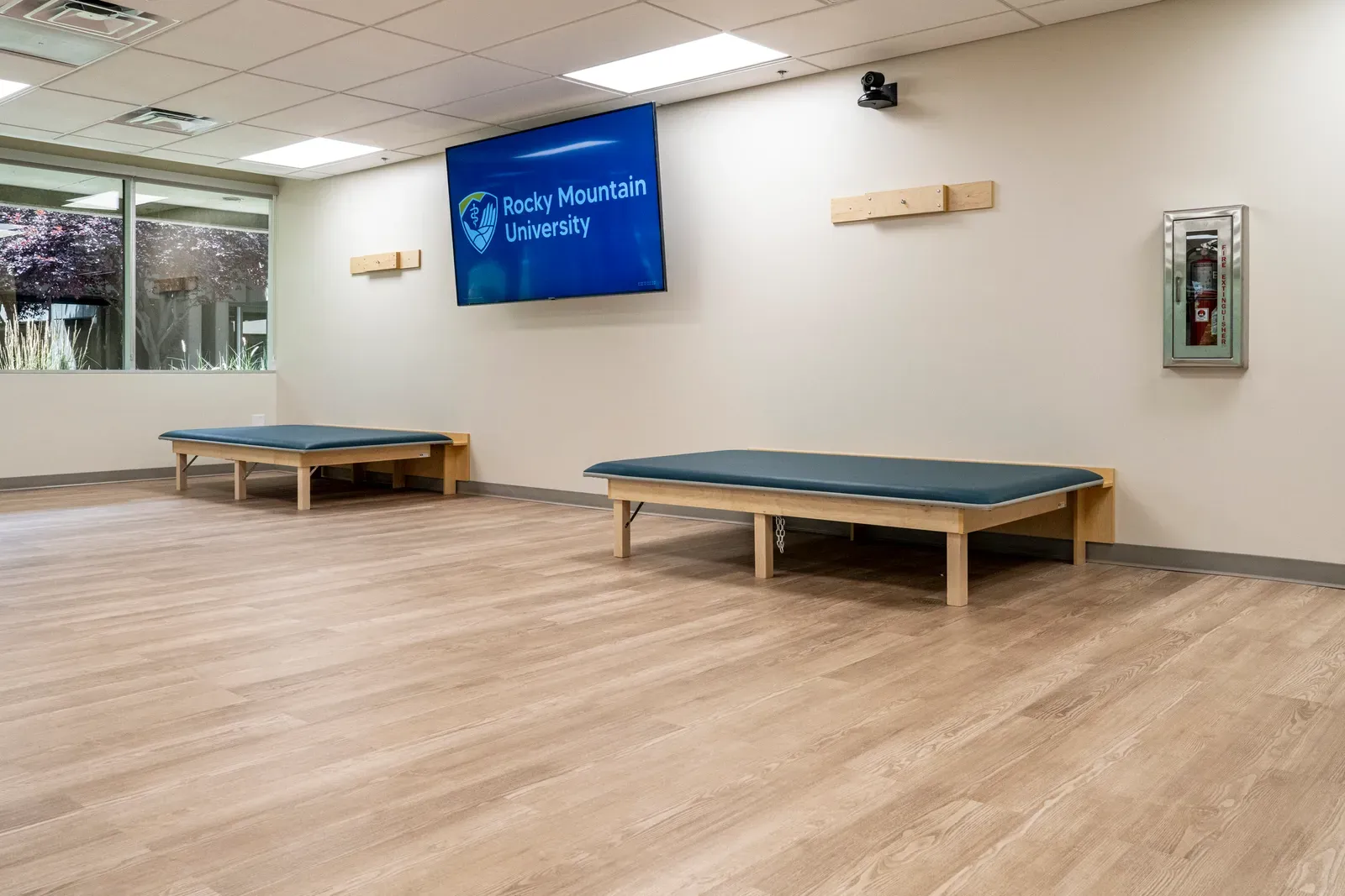 Therapy room with two low wooden treatment tables, a wall-mounted Rocky Mountain University screen and windows on the left.