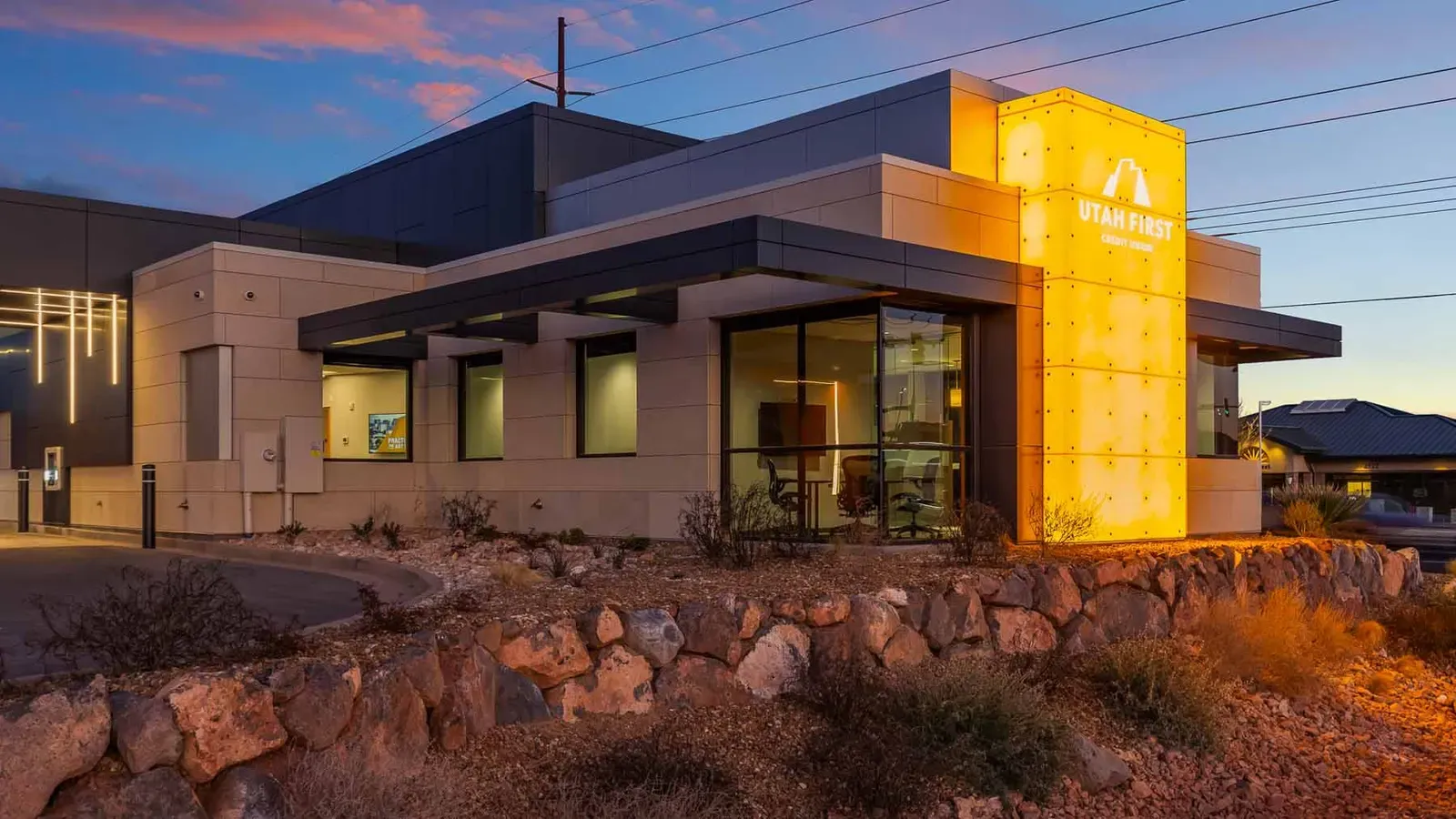 Exterior of the branch at dusk with a lit Utah First sign on a glowing panel and a rock-edged landscaped bed.