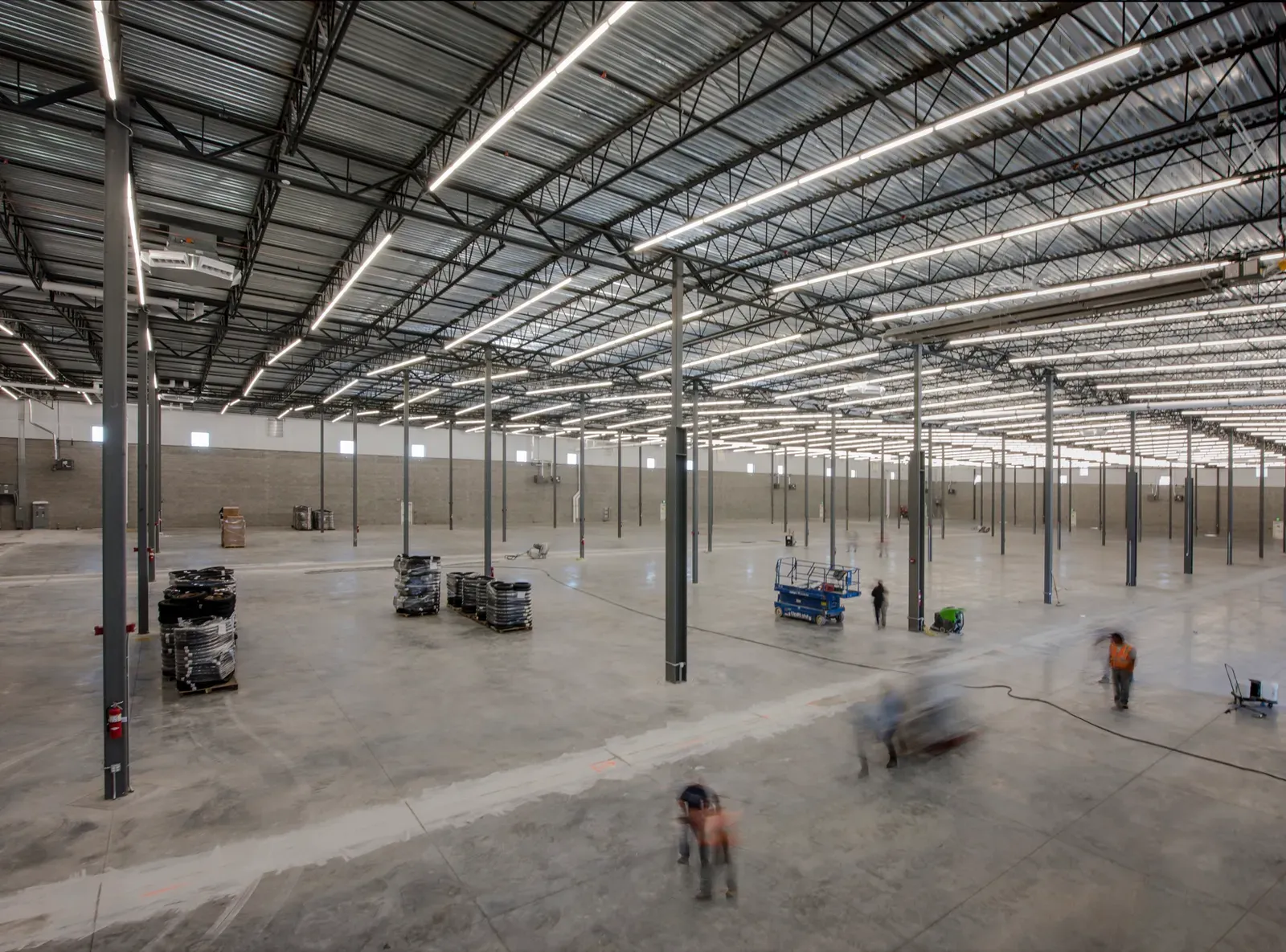 Open warehouse interior with steel columns, a steel roof structure and workers on a concrete floor.