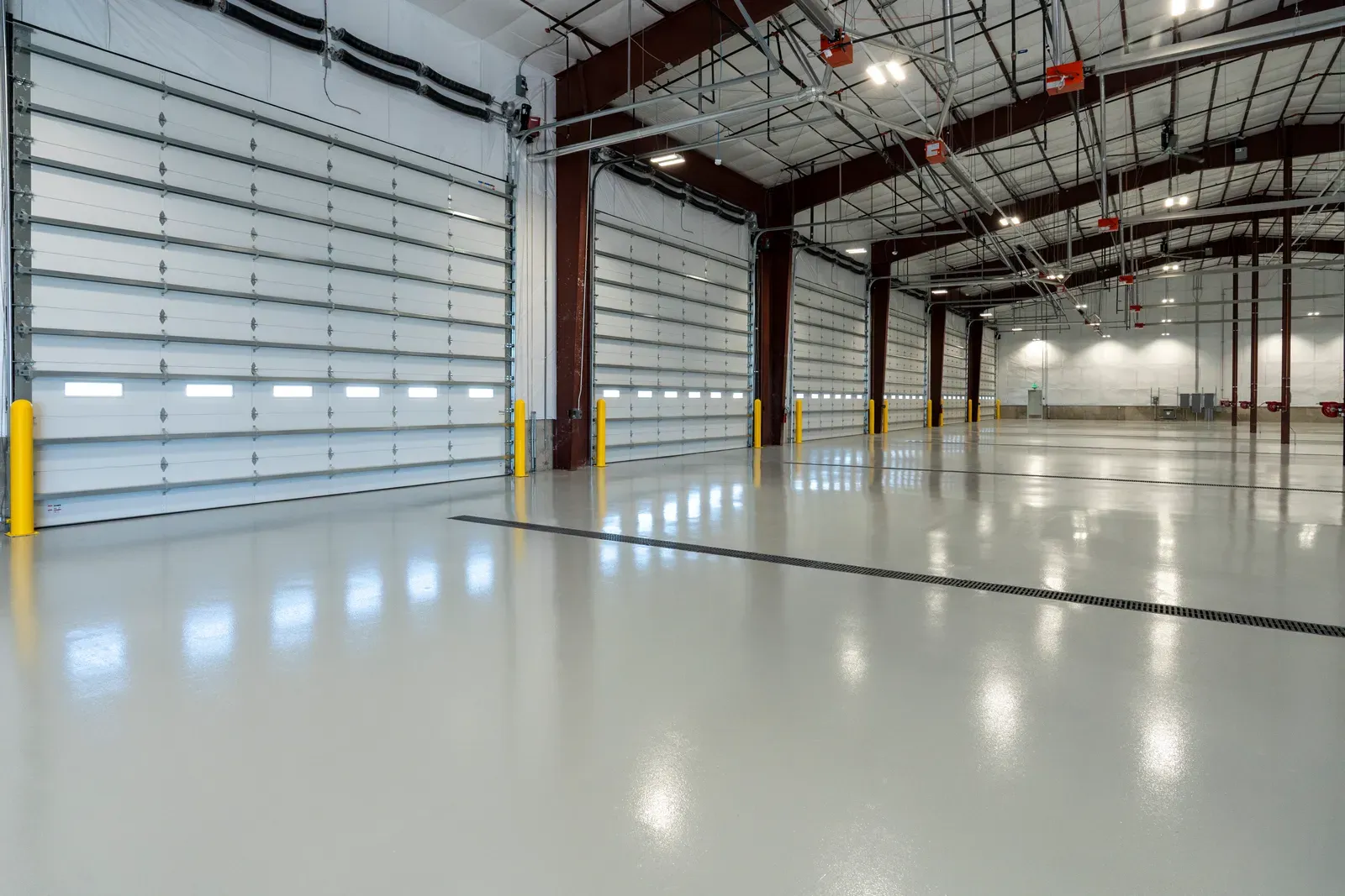Equipment storage bay with white overhead doors, red steel columns and a glossy gray floor with a trench drain.
