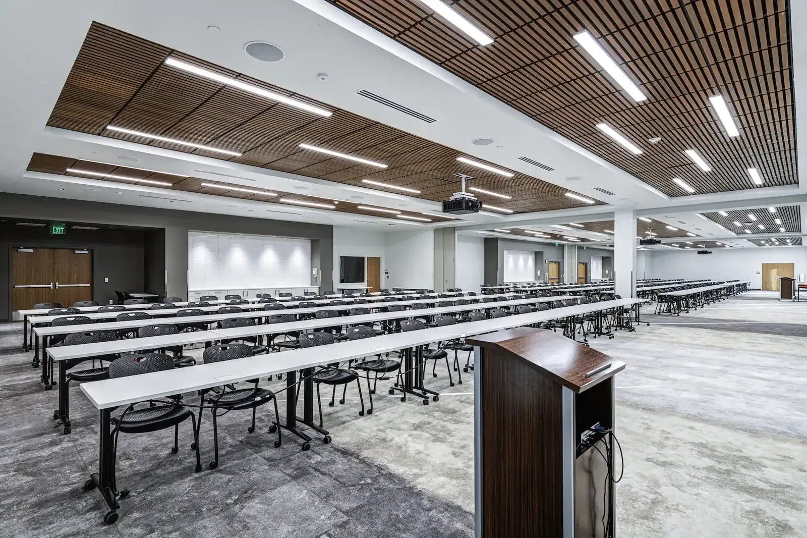 Large classroom with long rows of tables, a wood-slat ceiling, a projector and a podium.