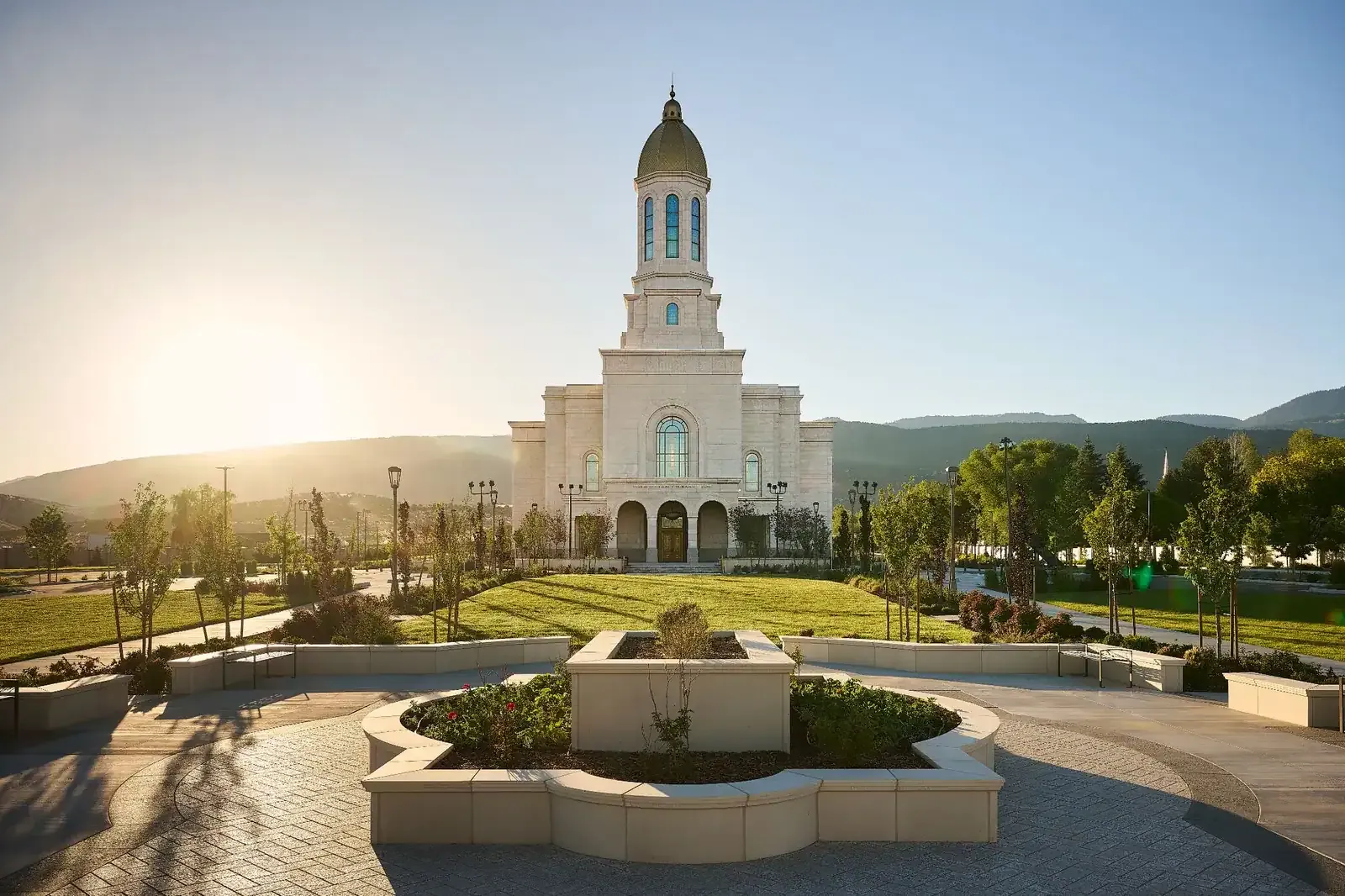 Front of the temple seen across a paved plaza with stone planters and landscaped lawns, with the sun low behind the hills