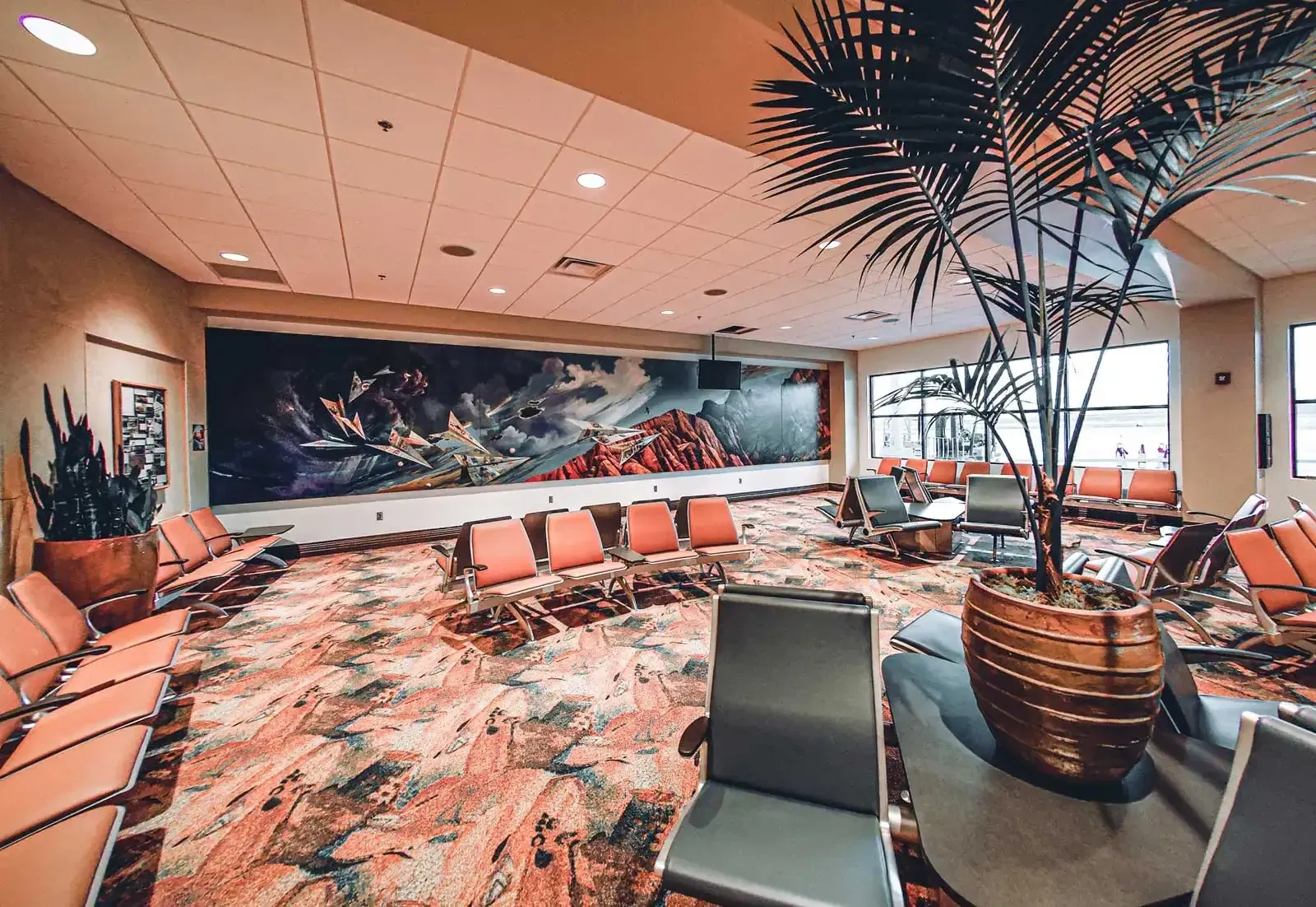 Gate seating area with orange chairs, patterned carpet, a large red-rock landscape mural on the wall and potted palms.