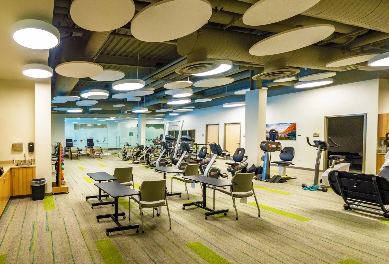 Rehabilitation gym with exercise machines, tables and green-striped carpet.