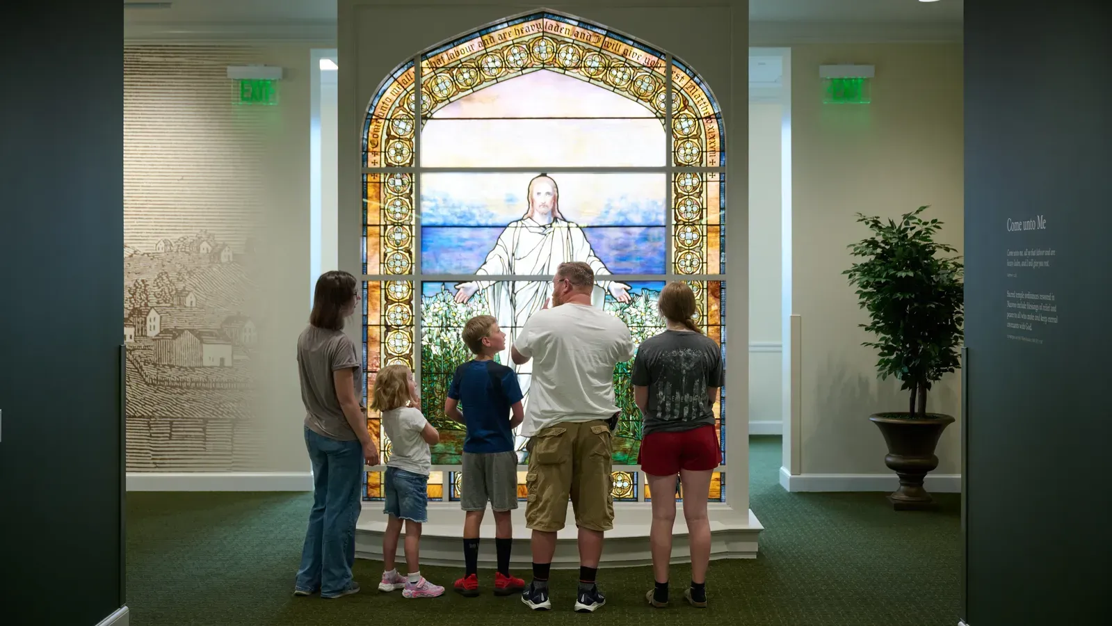 Visitors viewing a large arched art-glass window of Christ with outstretched arms in a green-carpeted gallery.